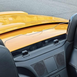 Può includere: Primo piano di un'auto cabriolet gialla con interni neri. L'immagine mostra il cruscotto, il parabrezza e una porzione del cofano dell'auto. L'auto è parcheggiata su una superficie di asfalto grigio.