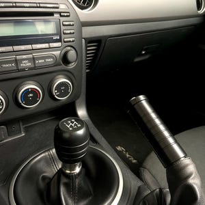 May include: Car interior showing a black gear shift knob with a 5-speed pattern and a black handbrake. The dashboard is grey and black, with air vents and a radio. The car's interior is dark, with leather and plastic components.