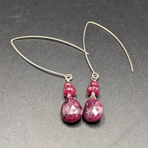 May include: A pair of silver-toned earrings featuring V-shaped ear wires. Each earring is adorned with a small faceted round ruby bead positioned above a larger faceted teardrop-shaped ruby bead, both secured with thin silver-toned wire wrapping.