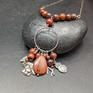 May include: A silver necklace featuring a pendant with a teardrop-shaped stone and various charms, including an owl, mushroom, and leaf. The necklace also has a strand of red and black speckled beads.