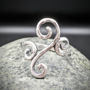 May include: A silver-toned ring with a hammered texture and a swirling, open design. The ring features three decorative swirls, creating a unique and artistic look. The ring is displayed on a gray stone surface.