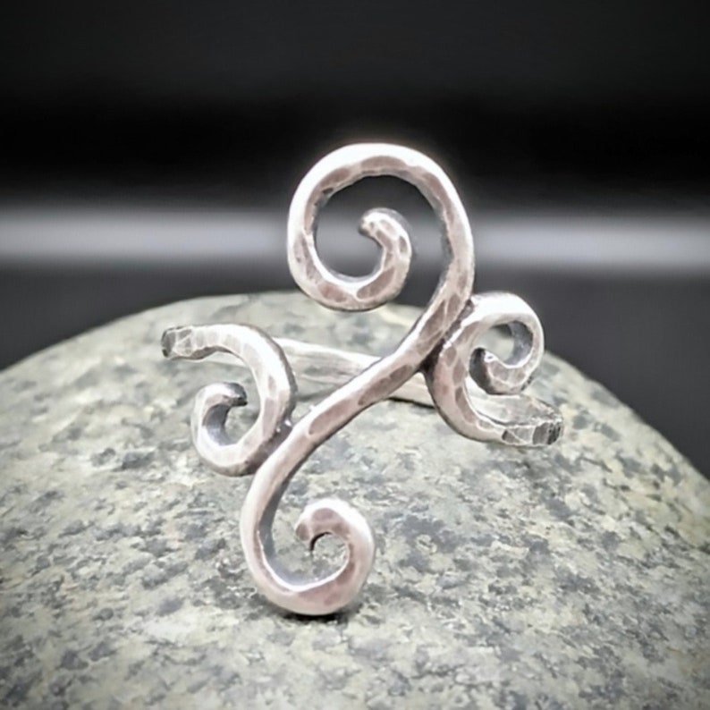 May include: A silver-toned ring with a hammered texture and a swirling, open design. The ring features three decorative swirls, creating a unique and artistic look. The ring is displayed on a gray stone surface.
