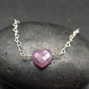May include: A silver chain necklace featuring a faceted, heart-shaped pink gemstone pendant. The pendant is a vibrant shade of pink, and the chain has a delicate, open link design. The necklace is displayed on a dark, textured surface.