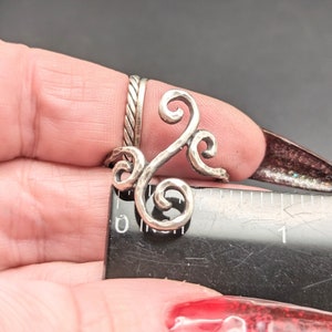 May include: A silver ring with a swirling, textured design. The ring is shown on a finger and measured against a ruler.