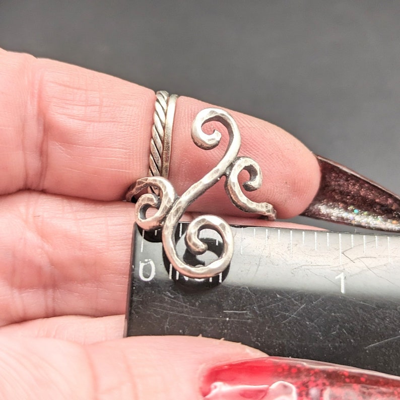May include: A silver ring with a swirling, textured design. The ring is shown on a finger and measured against a ruler.