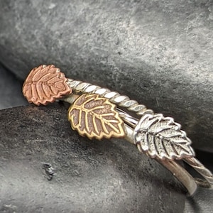 May include: Three silver rings with leaf designs. The top ring has a copper leaf, the middle ring has a gold leaf, and the bottom ring has a silver leaf.