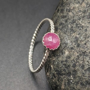 May include: A silver ring with a twisted band and a round, pink gemstone. The gemstone is set in a scalloped bezel. The ring is displayed on a dark gray surface.