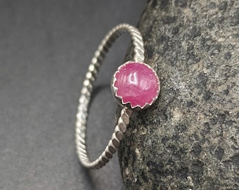 May include: A silver ring with a twisted band and a round, pink gemstone. The gemstone is set in a scalloped bezel. The ring is displayed on a dark gray surface.