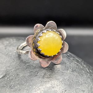 May include: A silver ring featuring a flower-shaped setting with a bright yellow gemstone. The ring's band has a textured design, and the gemstone is round and prominent, set against a dark background.