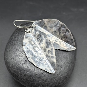 May include: A pair of silver leaf-shaped earrings. The earrings have a hammered texture and are suspended from silver ear wires. The leaves are slightly curved and have a naturalistic appearance, suggesting a botanical theme.