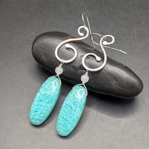 May include: A pair of silver-toned earrings with a unique, swirling design. Each earring features a light blue, elongated stone suspended below a small, round bead. The earrings are displayed on a dark, textured surface.