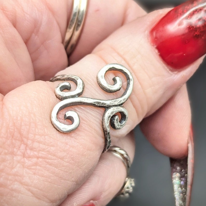 May include: A silver-toned ring with a unique, swirling design. The ring features three prominent spiral shapes connected by a horizontal bar. The metal has a hammered texture, giving it a rustic appearance. The ring is worn on a finger.