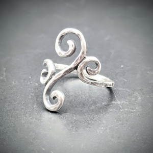 May include: A silver-toned ring with a hammered texture and a swirling, three-part design. The ring features three stylized curls, creating a unique and artistic look. The ring is set against a dark background.