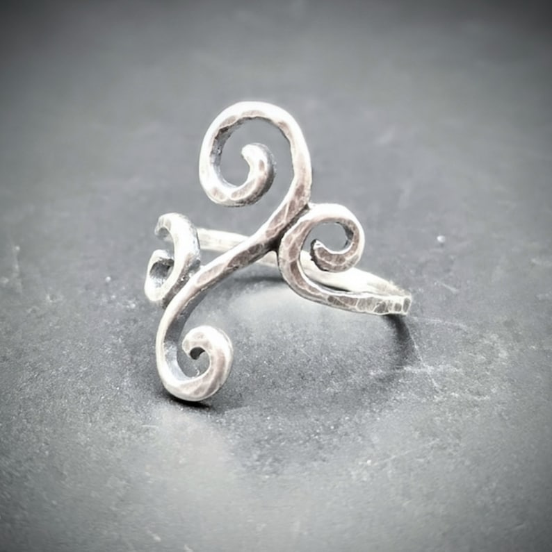 May include: A silver-toned ring with a hammered texture and a swirling, three-part design. The ring features three stylized curls, creating a unique and artistic look. The ring is set against a dark background.