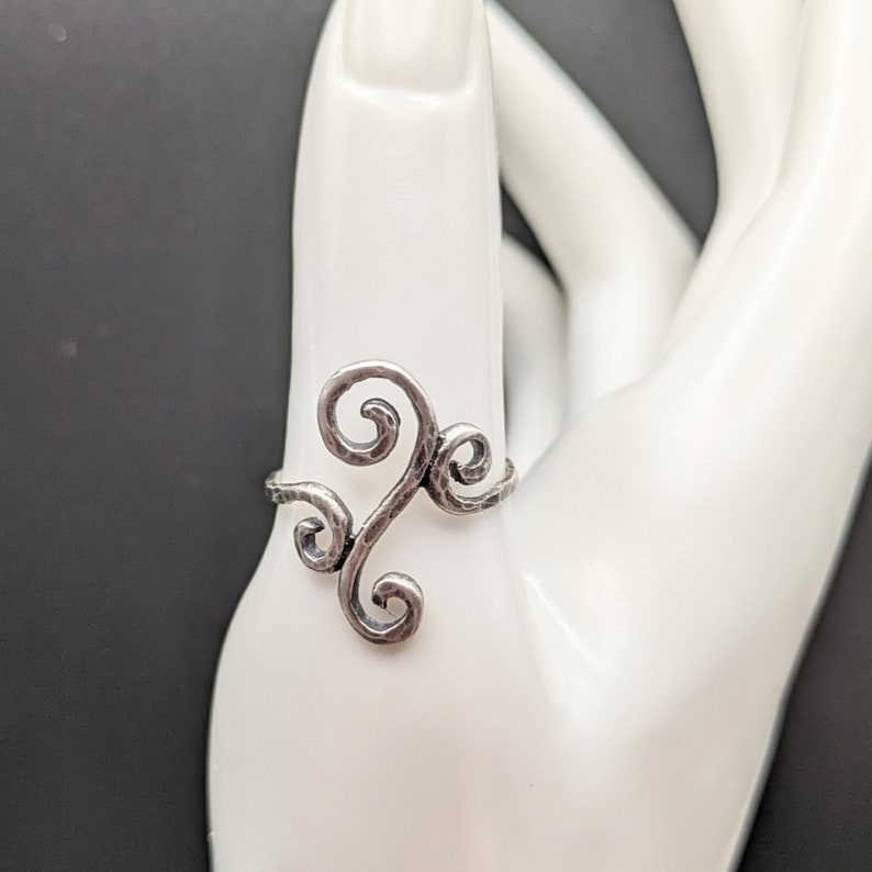 May include: A silver-toned ring featuring a textured, hammered metal surface with an openwork design composed of four distinct, interconnected swirling scroll motifs that extend vertically along the finger for a decorative appearance.