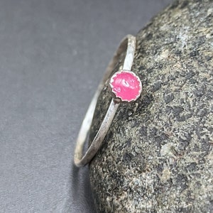 May include: A silver ring with a small, round, bright pink stone. The ring band has a hammered texture. The stone is set in a decorative bezel. The ring is displayed on a gray, textured surface.