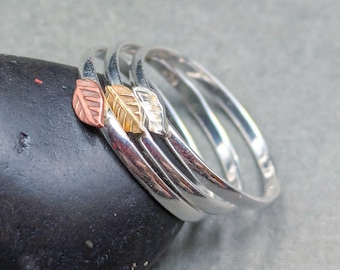May include: A set of three thin silver rings, each featuring a small metal leaf charm. One leaf is copper-toned, one is gold-toned, and one is silver-toned. The rings are stacked together, showcasing a polished metallic finish and delicate leaf details.