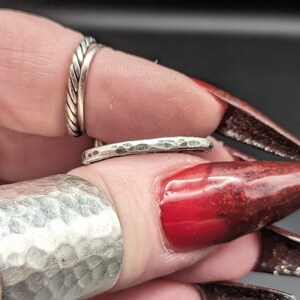 May include: A collection of silver rings, including a hammered band and a twisted rope design. The rings are displayed on a finger with red nail polish. The rings are likely made of sterling silver.
