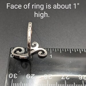 May include: A silver ring with a hammered texture and a decorative swirl design. The face of the ring is about 1 inch high. The ring is shown next to a ruler.