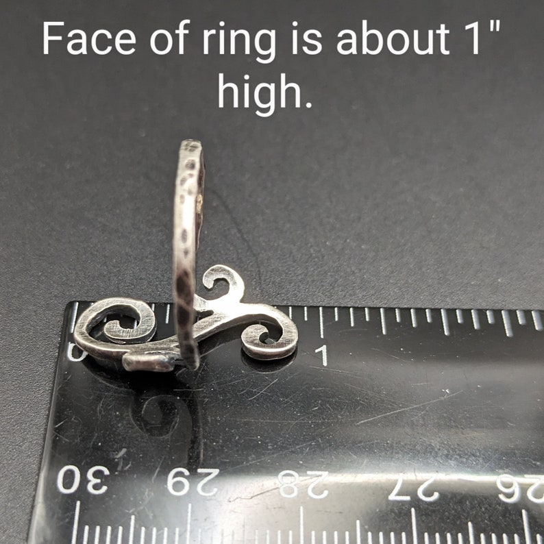 May include: A silver ring with a hammered texture and a decorative swirl design. The face of the ring is about 1 inch high. The ring is shown next to a ruler.