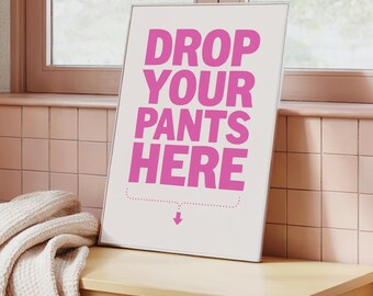 May include: A framed sign with the text "DROP YOUR PANTS HERE" in pink letters. The sign is on a light wood bench, with a cream-colored blanket draped over it. The background includes a window and pink tiled wall.
