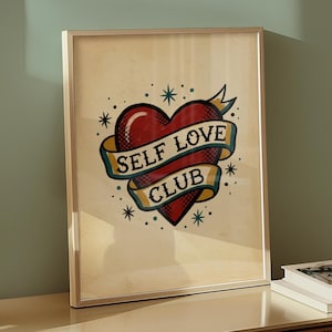 Può includere: Stampa artistica incorniciata con un cuore rosso e uno striscione con la scritta "SELF LOVE CLUB". Il design è in stile tatuaggio vintage, con stelle attorno al cuore. La cornice è in legno chiaro.