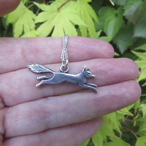 May include: A silver-toned metal pendant shaped like a running fox, attached to a delicate silver-toned chain. The fox is depicted in a horizontal, leaping pose with its tail extended behind it, featuring detailed textures on the fur and body.
