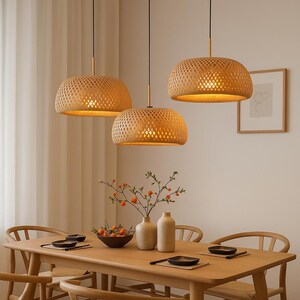 May include: Three woven pendant lights with a warm glow hang above a wooden dining table. The lampshades are a light brown color and have a textured, open weave design. The dining table is set with plates, bowls, and decorative items.