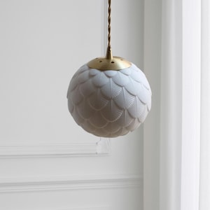 May include: A white, globe-shaped pendant light with a textured, feather-like pattern. The light fixture has a brass-colored top and a brown cord. The light hangs in front of a white wall and a white curtain.