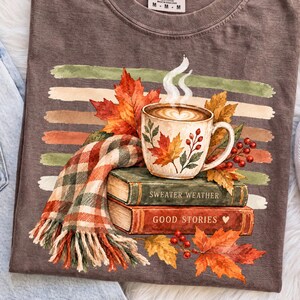 Puede incluir: Una camiseta marrón de manga corta con un gráfico de una taza de café humeante, una bufanda a cuadros y dos libros apilados con los títulos Sweater Weather y Good Stories, rodeados de hojas de otoño y bayas rojas sobre rayas horizontales.