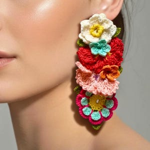 Pode incluir: Um par de brincos de flores de crochê vibrantes e feitos à mão. Os brincos apresentam uma cascata de flores coloridas em tons de branco, vermelho, rosa, laranja e turquesa, com centros amarelos e folhas verdes. Um acessório único e atraente.
