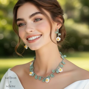 May include: A crochet necklace and earrings set featuring a floral design. The necklace and earrings are green with white and yellow flowers. The text "saffron + selene" is in the bottom left corner.