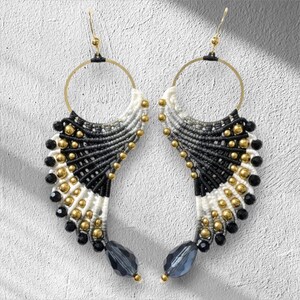 Pode incluir: Um par de brincos de macramé feitos à mão. Os brincos apresentam um design em forma de leque com fios pretos, brancos e cinzentos. Contas douradas e pretas adornam o design, com uma conta cinzenta em forma de gota na parte inferior.