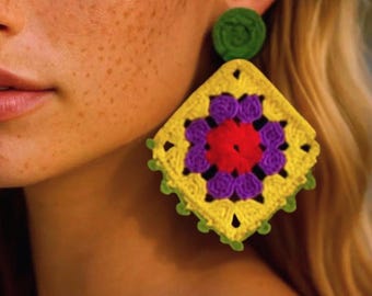May include: A close-up of a crochet earring. The earring features a square design with a yellow center, a red center, purple accents, and a green border. The earring is attached to a green circular stud.