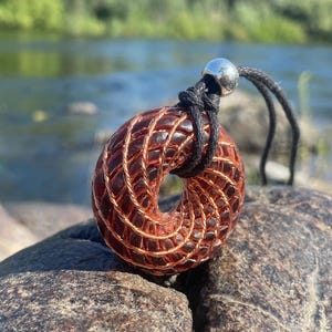 Könnte beinhalten: Halskette mit Anhänger in Donut-Form. Der rotbraune Resin-Anhänger hat Kupferdrahtdetails. Er hängt an einer schwarzen Kordel mit einer silbernen Perle. Die Halskette wird auf einem Stein präsentiert, mit einem Fluss im Hintergrund. Der Anhänger misst ca. 5 cm im Durchmesser.
