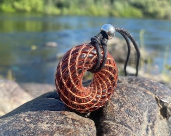 Puede incluir: Collar con colgante artesanal con un colgante de resina marrón rojizo en forma de rosquilla con detalles de alambre de cobre. El colgante está suspendido de un cordón negro con una cuenta plateada. El collar se exhibe sobre una roca, con un río al fondo.