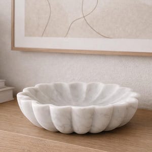 May include: A white marble bowl with a scalloped edge sits on a wooden shelf. Beside it are two small vases, a larger vase, and a decorative round object. A framed artwork hangs on the wall above the shelf, and a small vase of flowers sits on a stack of books.
