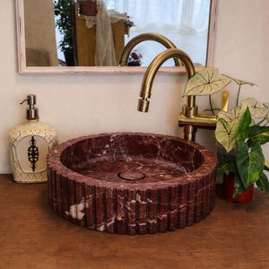 Peut inclure: Un &eacute;vier en marbre rouge avec un ext&eacute;rieur cannel&eacute; repose sur une surface en bois. Un robinet dor&eacute; et un distributeur de savon d&eacute;coratif sont visibles. Un miroir et de la verdure sont en arri&egrave;re-plan.