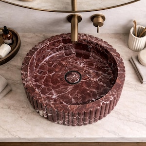 Peut inclure: Un lavabo de salle de bain rond en marbre rouge avec un ext&eacute;rieur cannel&eacute;. Le lavabo est encastr&eacute; dans un comptoir en marbre, avec un robinet et un miroir en laiton au-dessus. Accessoires : plateau en bois avec savon et flacons, peigne et serviettes.
