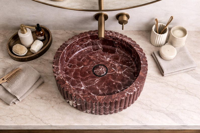 Peut inclure: Un lavabo de salle de bain rond en marbre rouge avec un ext&eacute;rieur cannel&eacute;. Le lavabo est encastr&eacute; dans un comptoir en marbre, avec un robinet et un miroir en laiton au-dessus. Accessoires : plateau en bois avec savon et flacons, peigne et serviettes.