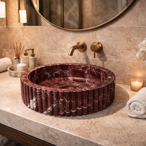 Peut inclure: Un &eacute;l&eacute;gant lavabo en marbre rouge avec un design cannel&eacute;. Le lavabo est pos&eacute; sur un comptoir en marbre, avec une robinetterie dor&eacute;e et un miroir. Des serviettes blanches roul&eacute;es, des bougies et des orchid&eacute;es compl&egrave;tent le d&eacute;cor.