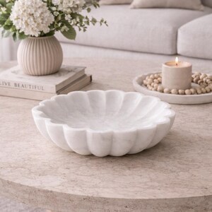 May include: A white marble bowl with a scalloped edge sits on a round, light-colored coffee table. A vase of white flowers and a lit candle are also on the table. The word "LIVE BEAUTIFUL" is visible on a book.
