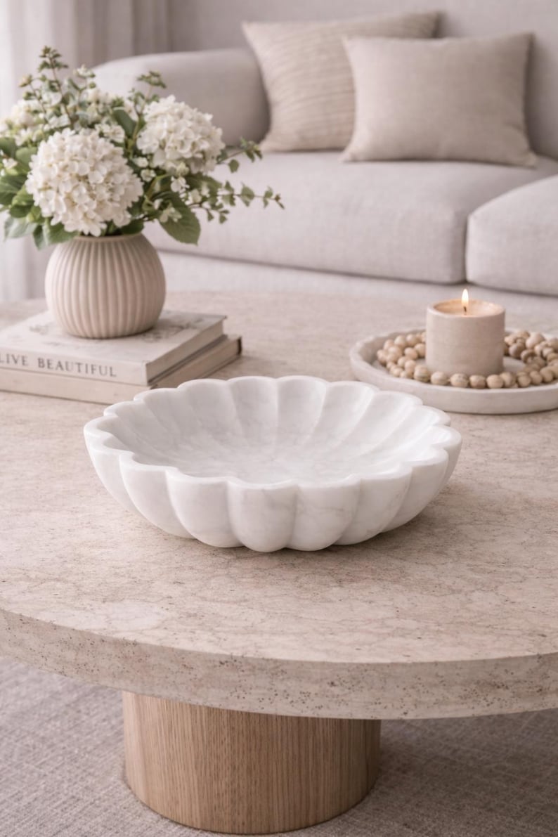 May include: A white marble bowl with a scalloped edge sits on a round, light-colored coffee table. A vase of white flowers and a lit candle are also on the table. The word "LIVE BEAUTIFUL" is visible on a book.