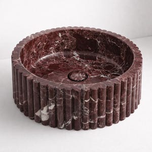 Peut inclure: Un &eacute;vier rond en marbre bordeaux avec un ext&eacute;rieur cannel&eacute;. L'&eacute;vier a un bassin circulaire et un drain au centre. Le marbre pr&eacute;sente des veines blanches. C'est un &eacute;l&eacute;ment de salle de bain.