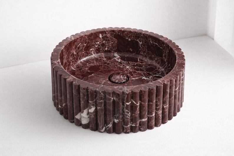 Peut inclure: Un &eacute;vier rond en marbre bordeaux avec un ext&eacute;rieur cannel&eacute;. L'&eacute;vier a un bassin circulaire et un drain au centre. Le marbre pr&eacute;sente des veines blanches. C'est un &eacute;l&eacute;ment de salle de bain.