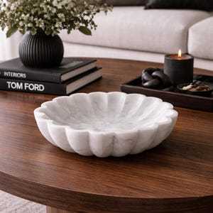 May include: A white marble decorative bowl with a scalloped edge sits on a dark wood coffee table. A black vase with flowers, books, and a candle are also on the table. The bowl is a decorative home accent.