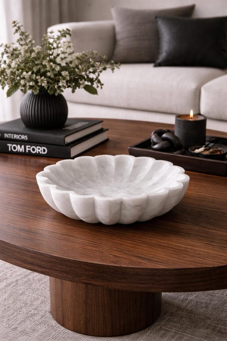 May include: A white marble decorative bowl with a scalloped edge sits on a dark wood coffee table. A black vase with flowers, books, and a candle are also on the table. The bowl is a decorative home accent.