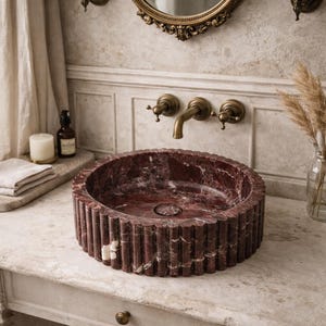 Peut inclure: Un &eacute;vier rond en marbre rouge avec un ext&eacute;rieur cannel&eacute; est pos&eacute; sur une vanit&eacute; en marbre. L'&eacute;vier a un drain central et est entour&eacute; de robinetterie en laiton, dont un robinet et deux appliques. Un miroir d&eacute;coratif est suspendu au-dessus.