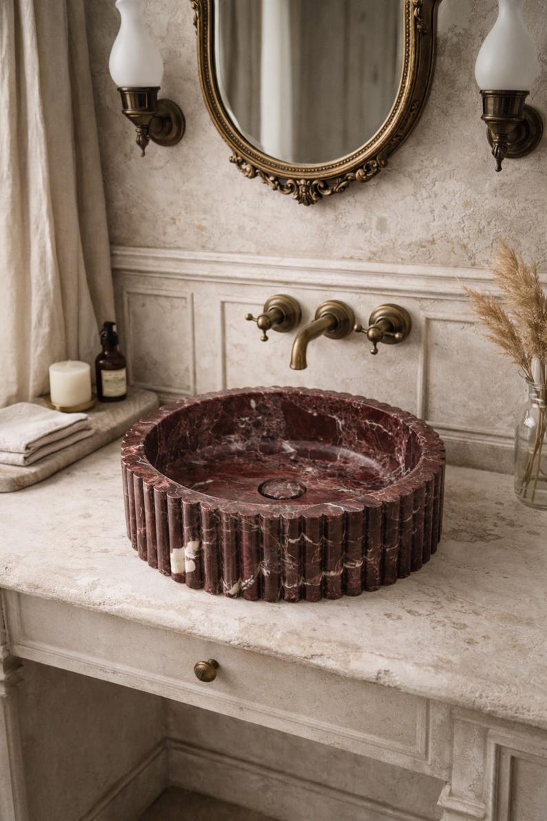 Peut inclure: Un &eacute;vier rond en marbre rouge avec un ext&eacute;rieur cannel&eacute; est pos&eacute; sur une vanit&eacute; en marbre. L'&eacute;vier a un drain central et est entour&eacute; de robinetterie en laiton, dont un robinet et deux appliques. Un miroir d&eacute;coratif est suspendu au-dessus.