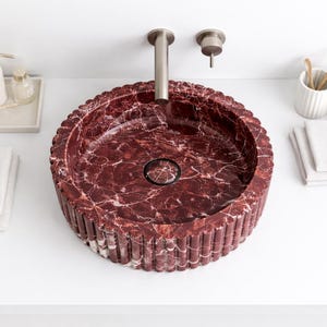 Peut inclure: Un lavabo rond en marbre bordeaux avec un ext&eacute;rieur cannel&eacute;. Le lavabo est pos&eacute; sur une surface blanche, avec un robinet argent&eacute; et d'autres accessoires de salle de bain, dont un distributeur de savon, du savon et des serviettes. L'int&eacute;rieur du lavabo pr&eacute;sente un motif marbr&eacute;.